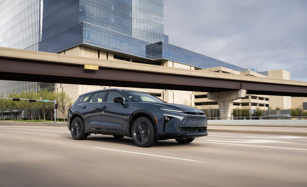 front three quarter view of a dark gray suv driving on a highway with modern buildings in the background.